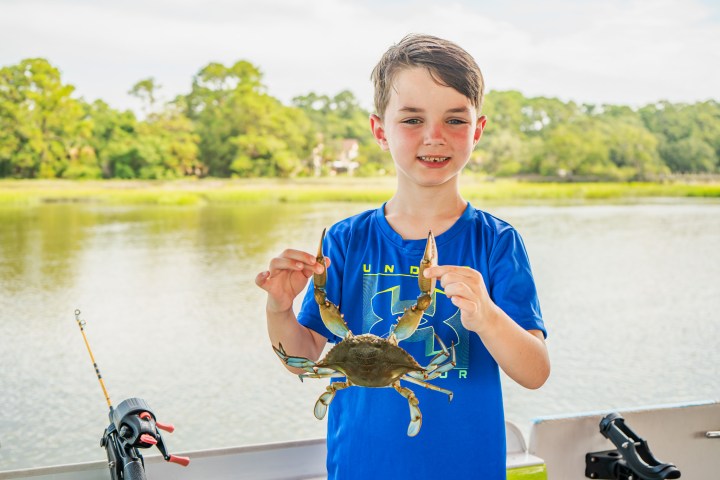 Young boy in blue shirt holding a crab near a lake with fishing rods visible.