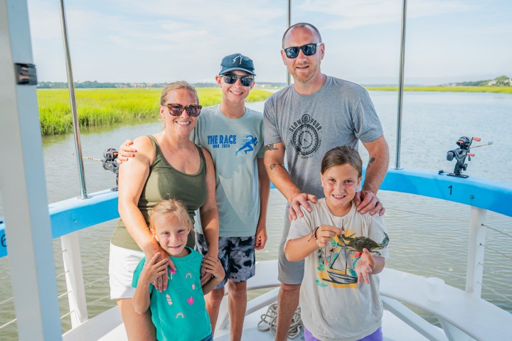 Family of five on a boat, smiling with a child holding a crab, water and greenery background.