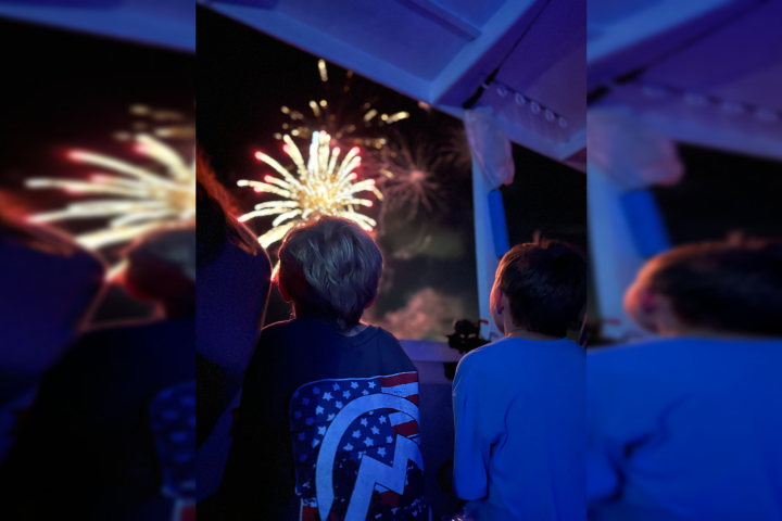 Two children watching colorful fireworks through a window at night.