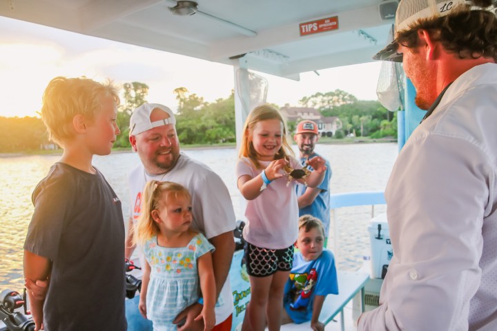Children on a boat smiling and holding a small crab, with adults watching.